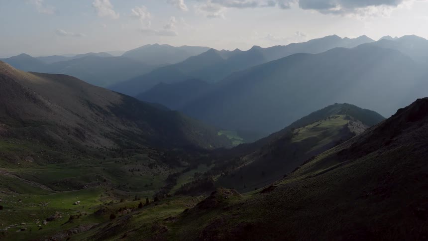 A panoramic drone shot glides over a deep mountain valley revealing silhouettes of jagged peaks that stretch toward a hazy horizon under a bright daytime sky
