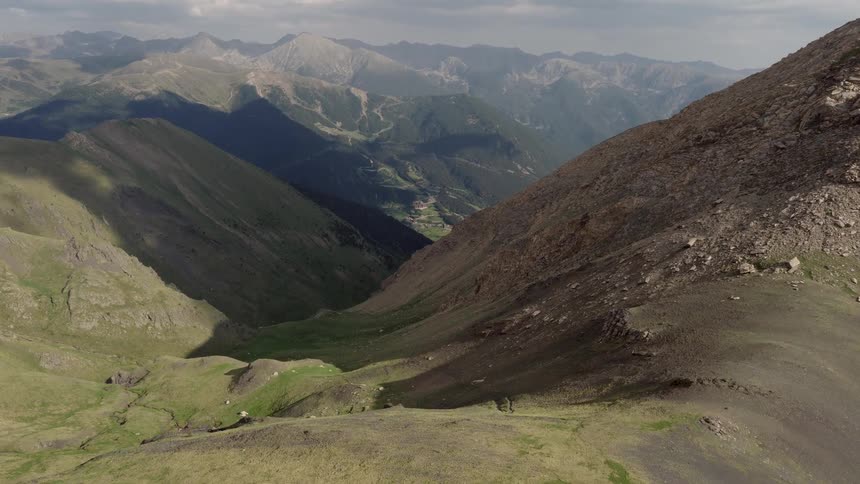 A view from above showing green and rocky mountain peaks stretching toward a distant valley under a cloudy sky during the day