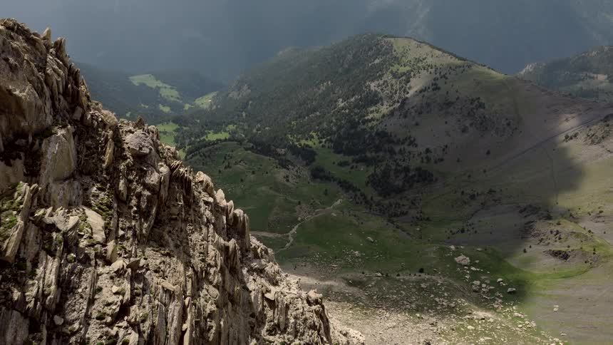 An aerial shot captures a steep rocky mountain peak overlooking a lush green valley nestled between distant slopesas the camera glides along the jagged ridge a dark bird flies across the vast sundrenched landscape