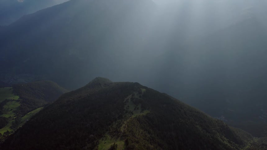 An aerial view captures layered mountain ridges fading into the distance with soft light filtering through the sky above a green hilltop in the foreground