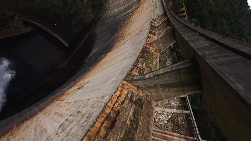 A drone dives rapidly down the curved concrete face of a massive dam toward a powerful white water spray discharging into the dark valley below while steep forested mountains and a cloudy sky remain visible in the background of the shot