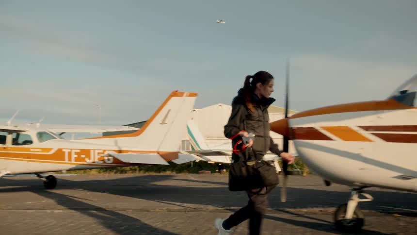 A woman carrying a bag walks across a paved area toward a small white and orange airplane parked under a clear sky during the day