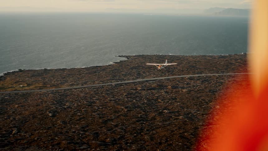 An aerial view shows a plane navigating a stark landscape near the sea