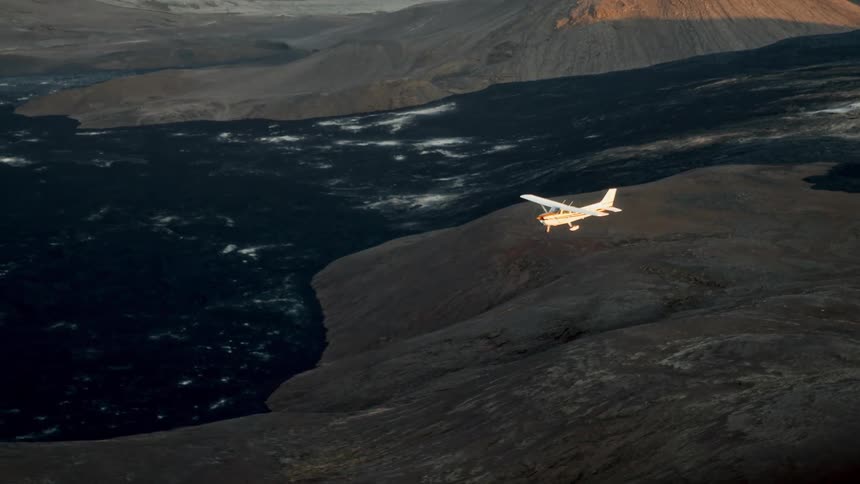 A small white airplane flies over a vast landscape of dark volcanic rock and hills during the late afternoon