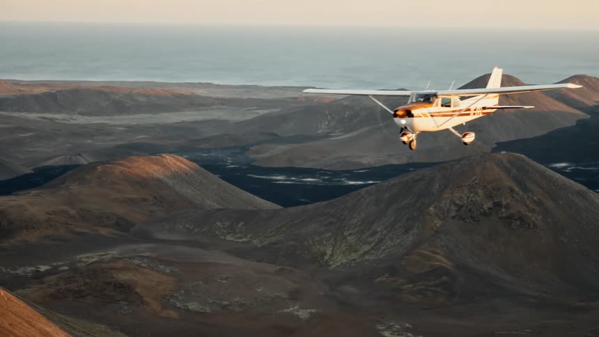 A small orangeandwhite propeller plane flies above rugged brown mountains with the ocean visible in the distance under a clear sky