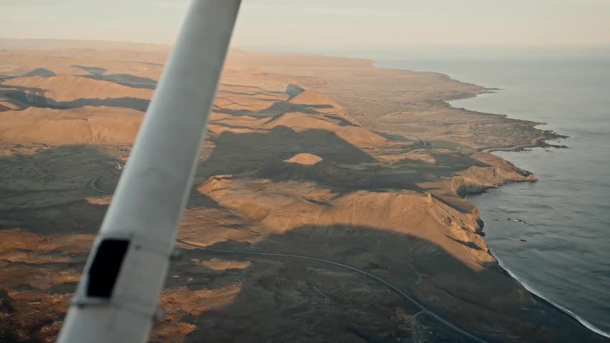 An aerial perspective from a small plane flying over a rugged sundrenched coastline at golden hour it captures the dramatic contrast between the rolling desert hills and the deep blue ocean as a road winds along the shore below