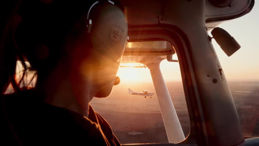 A female pilot wearing a headset flies a plane during sunset while another small aircraft is visible through the side window against a golden horizon