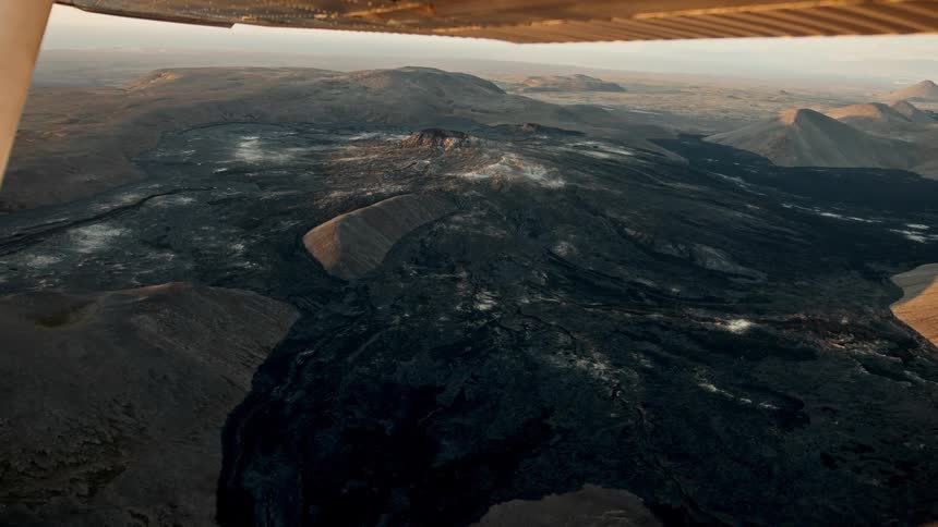 An aerial view from a small plane reveals a vast dark volcanic landscape filled with hardened lava flows and craters the golden hour light casts long shadows over the rugged terrain highlighting steam rising from the volcanic vents in the distance