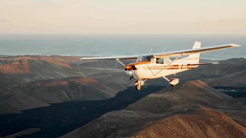 A small orangeandwhite propeller plane flies above rugged brown mountains with the ocean visible in the distance under a clear sky