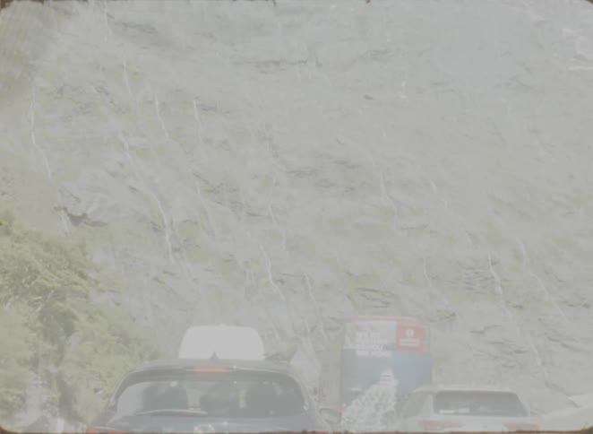 A line of cars stopped on a road directly beneath a steep rocky cliffside rain or mist falls across the scene creating a hazy atmosphere as the vehicles wait near the base of the mountain