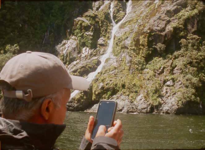 Man photographs a waterfall with a phone by a lake surrounded by rocks