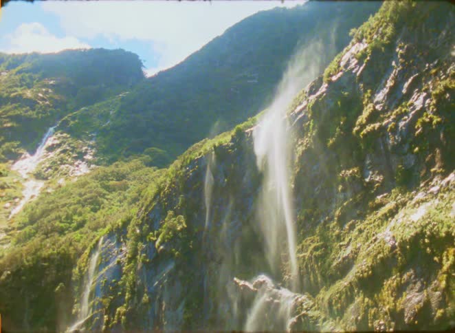 A view of a thin waterfall cascading down a steep rocky cliff covered in green plants and moss on a sunny day