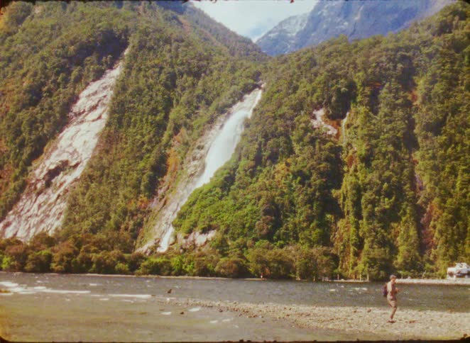 Man walks along a riverbank with a waterfall between vegetationcovered mountains