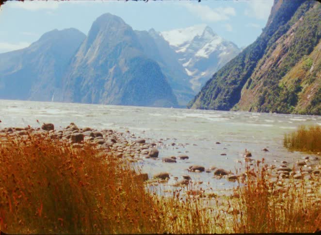 A wide glacial lake surrounded by massive steep mountains and a snowy peak under a bright sky while brown tall grass and smooth rocks sit in the foreground bank