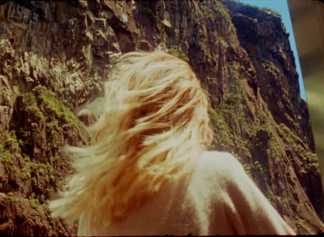 A young blonde woman standing beside a rock in the wild with the wind blowing through her long hair on a sunny day