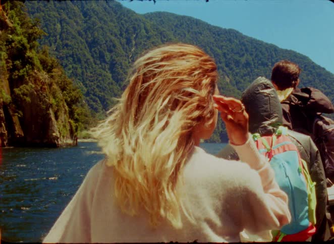 Woman on a boat adjusts her hair in the wind facing mountains and a lake