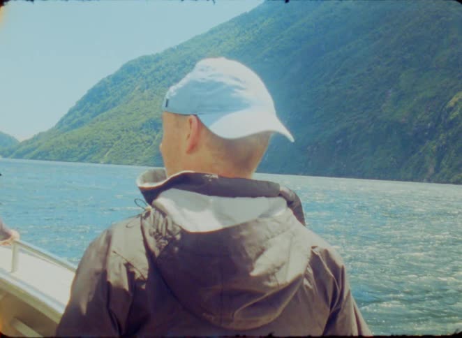 Man wearing a cap looks at a lake surrounded by green mountains