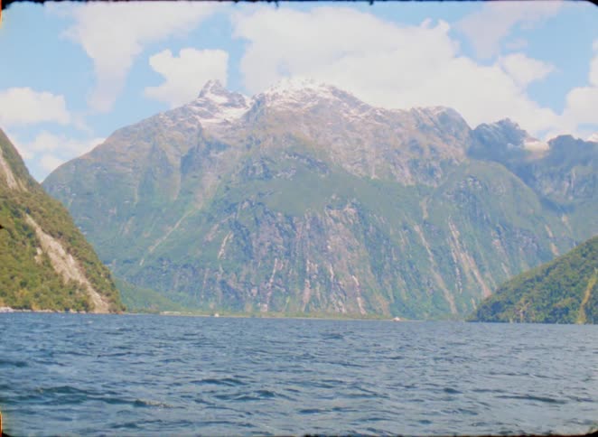 The camera captures a wide view of a massive mountain rising steeply from the deep blue waters of a fjord under a bright sky with scattered clouds and patches of snow visible on the high rocky peaks in the distance