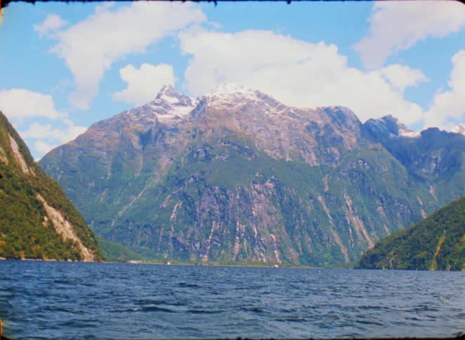 A view from a boat shows dark blue water in the foreground with massive green mountains and a snow capped peak under a blue sky with white clouds