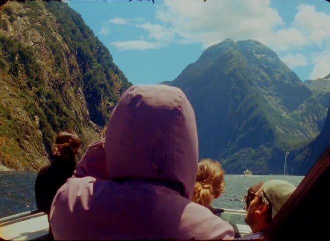 A group of people sits on a boat as it travels through a deep blue fjord surrounded by steep green mountains under a bright sky while a small waterfall and another vessel appear in the distant background of the mountainous landscape