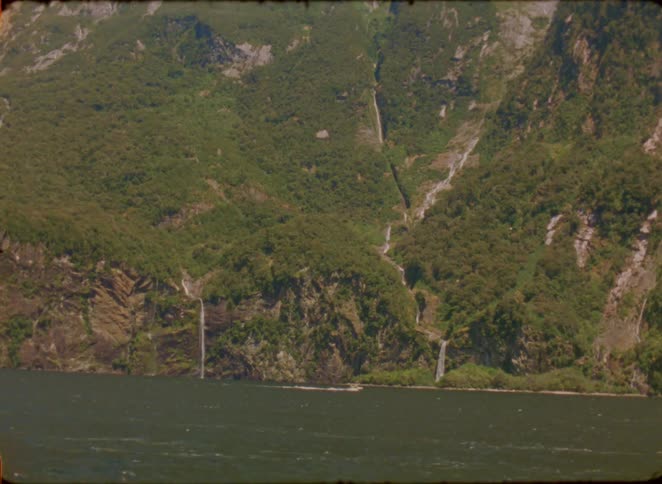 A wide shot captures a boat traveling across a large body of water toward a massive mountain range where several waterfalls cascade down steep green cliffs into the sea under a clear sky in a remote and natural wilderness setting