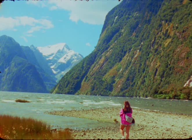 A woman in a pink shirt stands on a rocky shore looking out toward a turquoise bay framed by towering green cliffs in the distance a majestic snowcapped mountain peak rises sharply against a clear blue sky