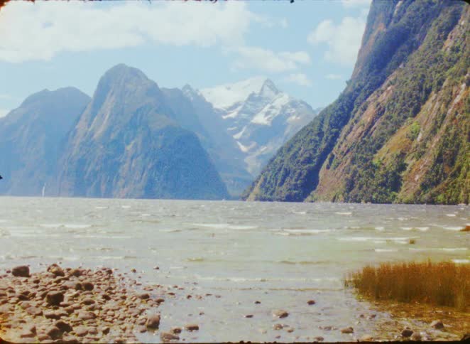 A view shows a wide blue lake with small waves near a rocky shore under a blue sky with white clouds while massive green mountains and a snow capped peak tower in the distance