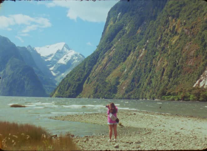 A woman in a pink shirt and white shorts stands on a rocky shore and looks toward massive green mountains and a snow capped peak under a blue sky during the day