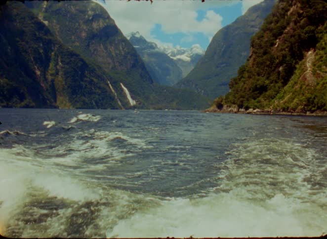 A view from a boat shows churning white wake in the foreground with massive green mountains and a snow capped peak under a blue sky with white clouds