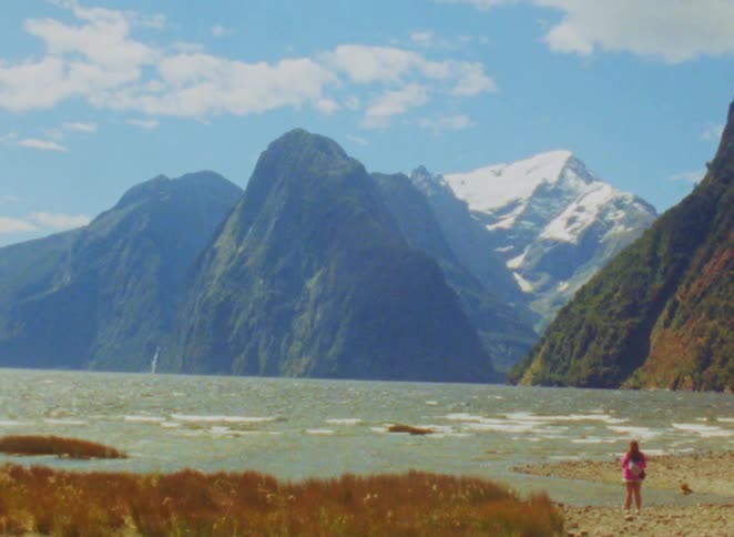 Person walking along a lakeshore with mountains and snowcapped peaks in the background
