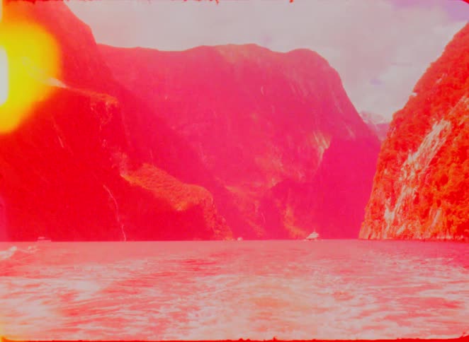 A scenic boat journey through a deep mountain fjord featuring steep green cliffs and a wake trailing in the water as the clip progresses it is overtaken by heavy vintage film burns and orange light leaks that eventually wash out the entire frame