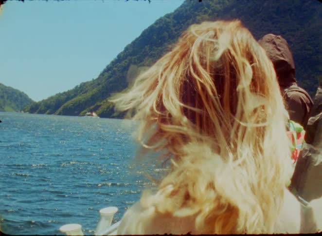 A woman with blonde hair stands on a boat as the wind blows her hair while looking out at deep blue water and lush green mountains under a bright sky during the day
