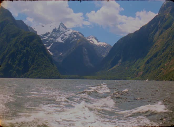 A boat moves through a deep fjord leaving a white wake on the water while massive snow capped mountains and green slopes rise toward a blue sky with fluffy clouds