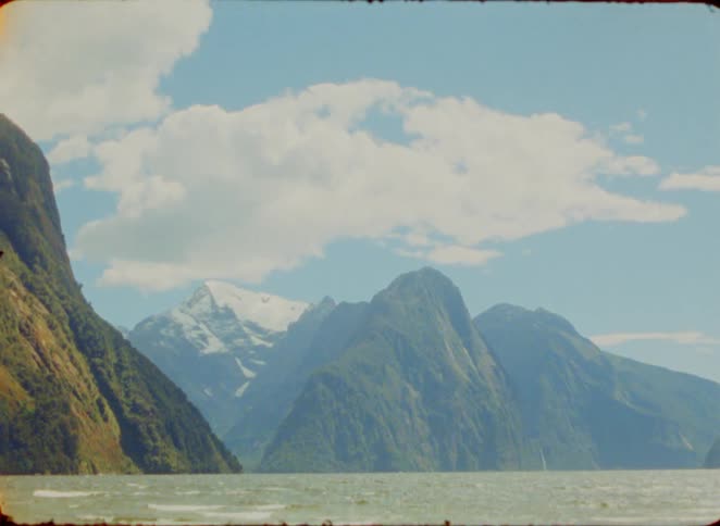Lake surrounded by green mountains with snowcapped peaks under a partly cloudy sky