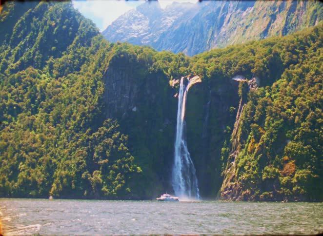 A tall slender waterfall plunges down a lush green cliffside into a body of water where a small boat is positioned near the base steep forested mountains rise in the background under a bright sky framing the dramatic coastal landscape
