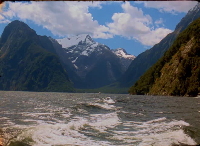 A scenic view from the back of a boat as it cruises through a deep blue fjord surrounded by towering lush green mountains in the distance a prominent snowcapped peak stands out against a bright sky filled with soft white clouds