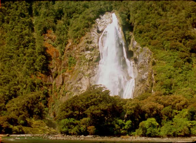 A powerful waterfall cascades down a lush green mountain slope into a valley surrounded by dense forest under a clear sky during the day