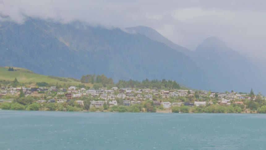 A view of many small white houses along a green shore next to a large body of blue water with tall mountains covered in mist in the background