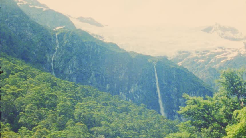 Waterfalls flow down green mountains with snowy areas in the background