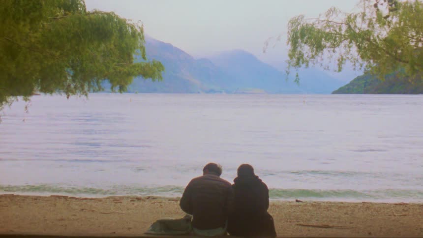 Couple sitting on a lakeshore with mountains in the background and trees framing the landscape