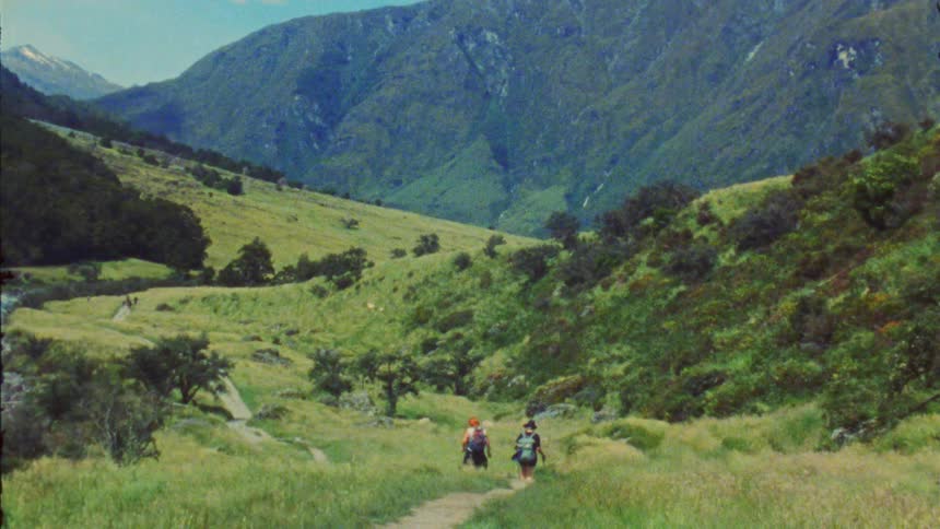 Two people hiking down a hill in the wilderness surrounded by a mountain range covered in green vegetation at midday
