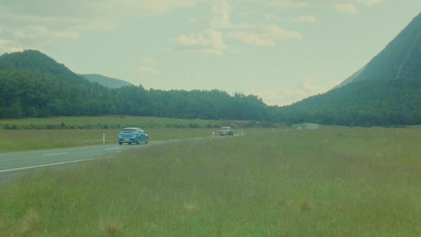 Multiple cars drive along a long road through a vast green valley between lush hills under a blue sky with white clouds during the day