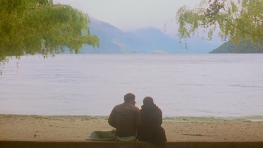 Couple sitting on the sand looks at a lake with mountains in the background