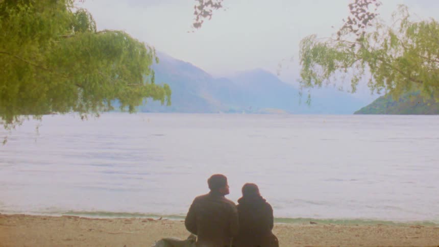 Young adult man and woman sitting on a beach next to the sea with a mountain range on the other side of the shore outdoors during day time