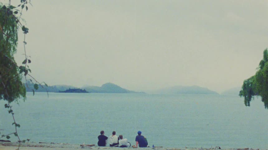 A group of friends sits on a sandy beach looking out at a wide blue lake with distant islands under a cloudy sky during the day