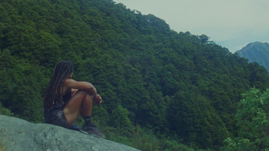A young woman sitting on a rock looking out at the vast forest in front of her at midday
