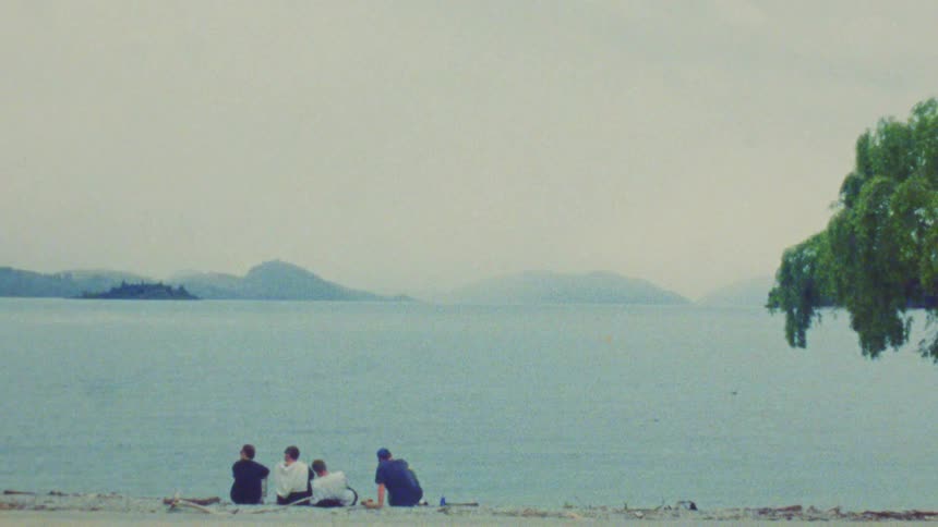 Four people sit on the sand by a lake with islands in the background