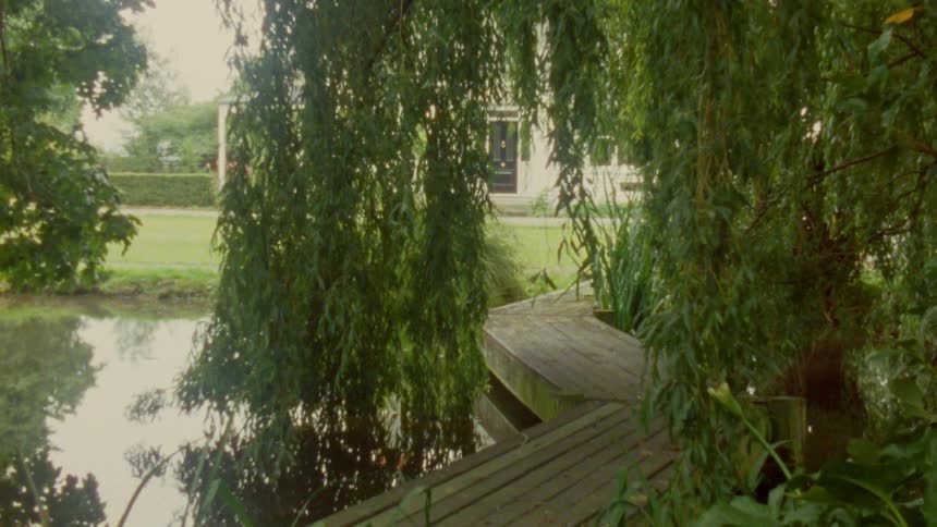 A walkway over a pond with trees behind a building at midday