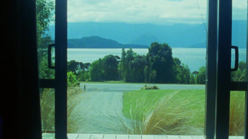 A view through open glass doors reveals a wooden deck and a gravel driveway leading toward green trees and a large body of water with blue mountains under a cloudy sky