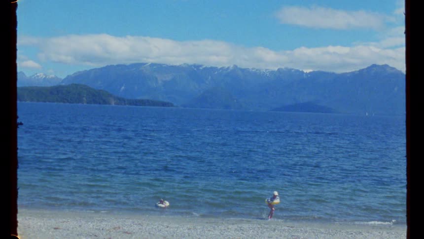 A child runs out of a lake after swimming holding a pool paddle while another child is still swimming at midday